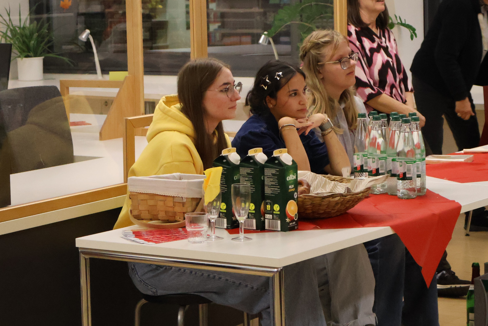 Drei Schülerinnen sitzen an einem Tisch mit Getränken und Brezeln, die sie während der Lesung in der Mediathek anbieten. Drei Schülerinnen sitzen an einem Tisch mit Getränken und Brezeln, die sie während der Lesung in der Mediathek anbieten.
