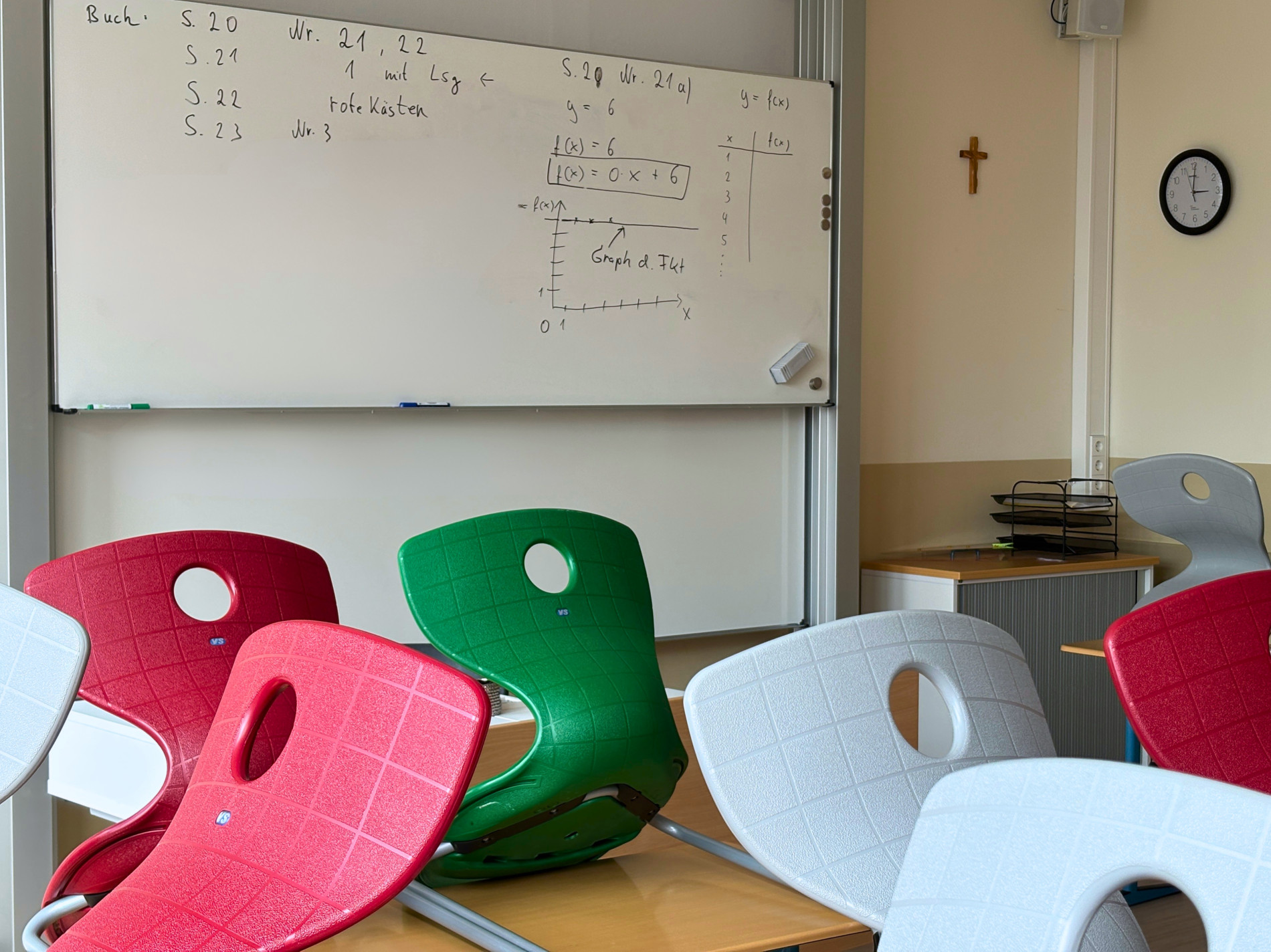 Klassenraum mit Tafel und bunten Stühlen, an der Wand hängt eines der neuen Kreuze aus Olivenholz.