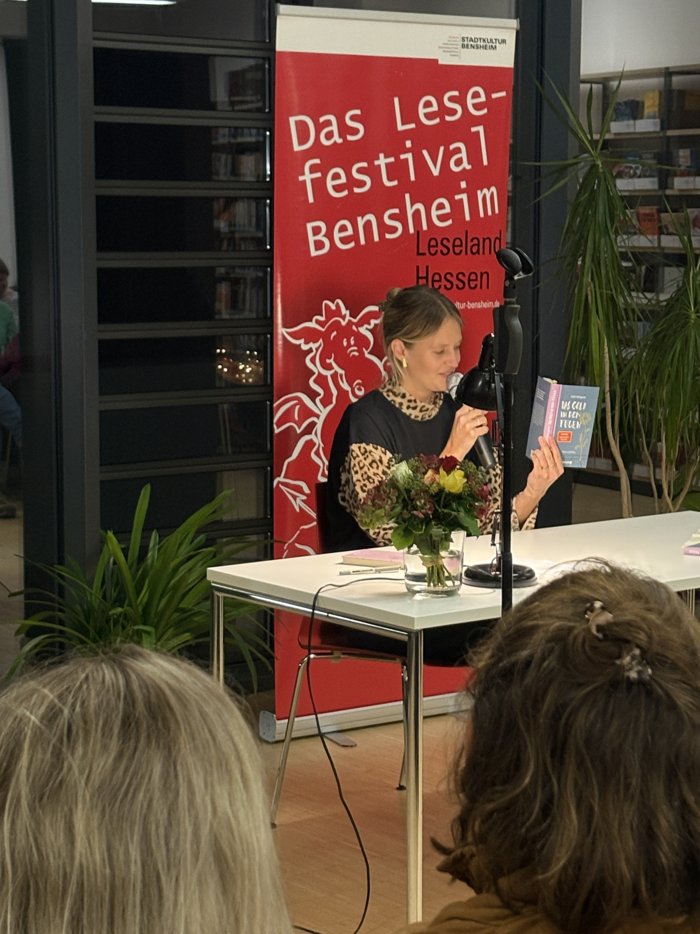 Leah Weigand sitzt am Lesungstisch, hält ihr Buch „Das Gold in den Fugen“ hoch und spricht in ein Mikrofon. Hinter ihr steht das Banner des Bensheimer Lesefestivals. Leah Weigand sitzt am Lesungstisch, hält ihr Buch „Das Gold in den Fugen“ hoch und spricht in ein Mikrofon. Hinter ihr steht das Banner des Bensheimer Lesefestivals.