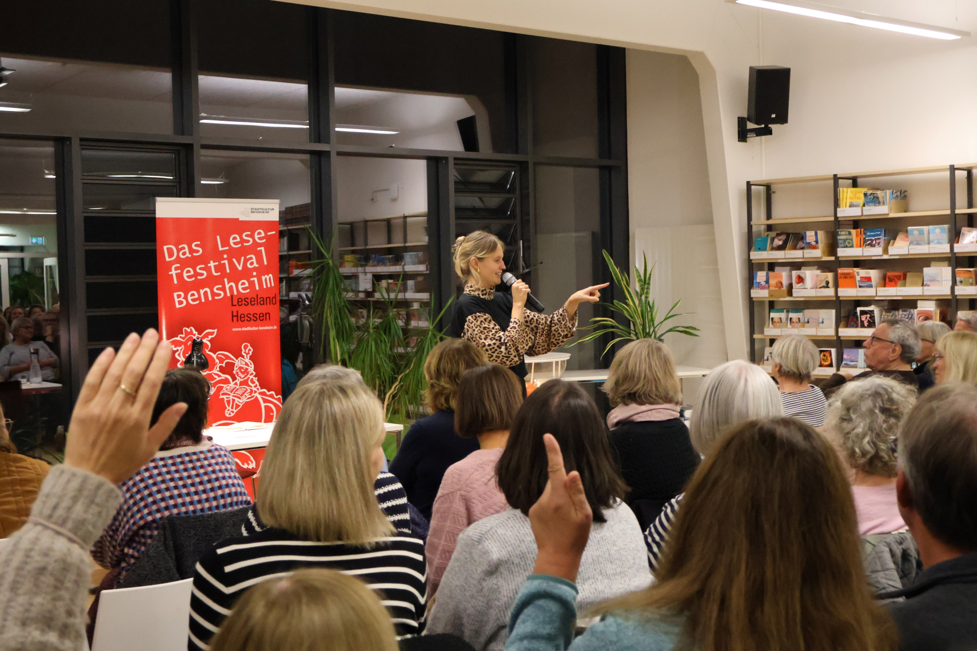 Leah Weigand spricht mit einem Mikrofon zum Publikum und zeigt mit der Hand in Richtung der Gäste. Die Stuhlreihen sind gut besetzt. Leah Weigand spricht mit einem Mikrofon zum Publikum und zeigt mit der Hand in Richtung der Gäste. Die Stuhlreihen sind gut besetzt.